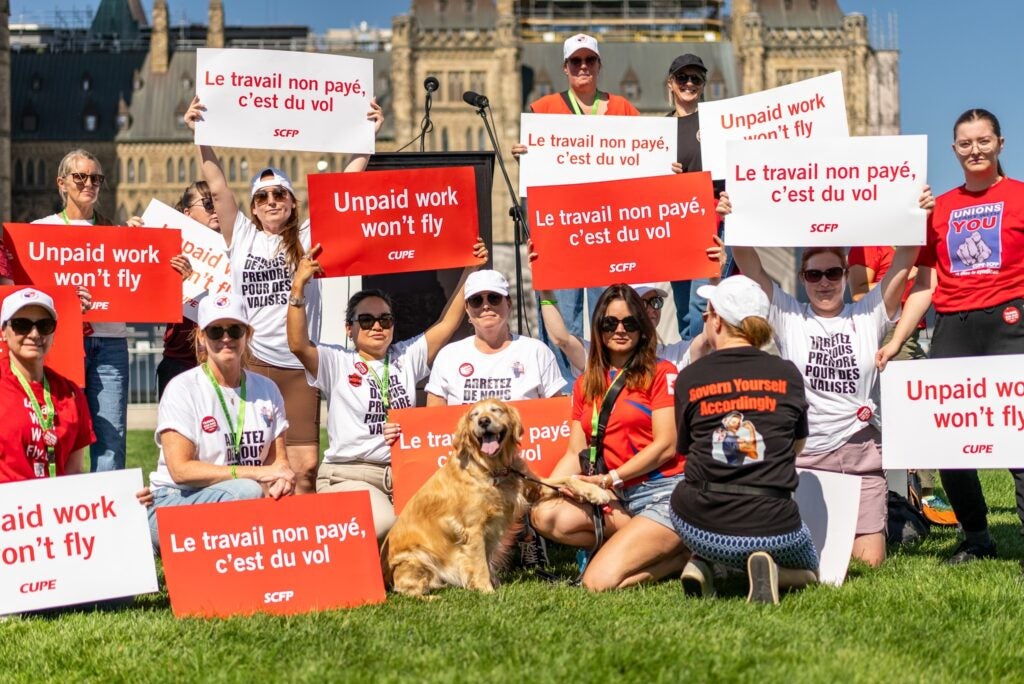 Numerous people sitting on the lawn in front of Parliament Hill holding "Unpaid work won't fly" and "Le travail non payé c'est du vol" signs