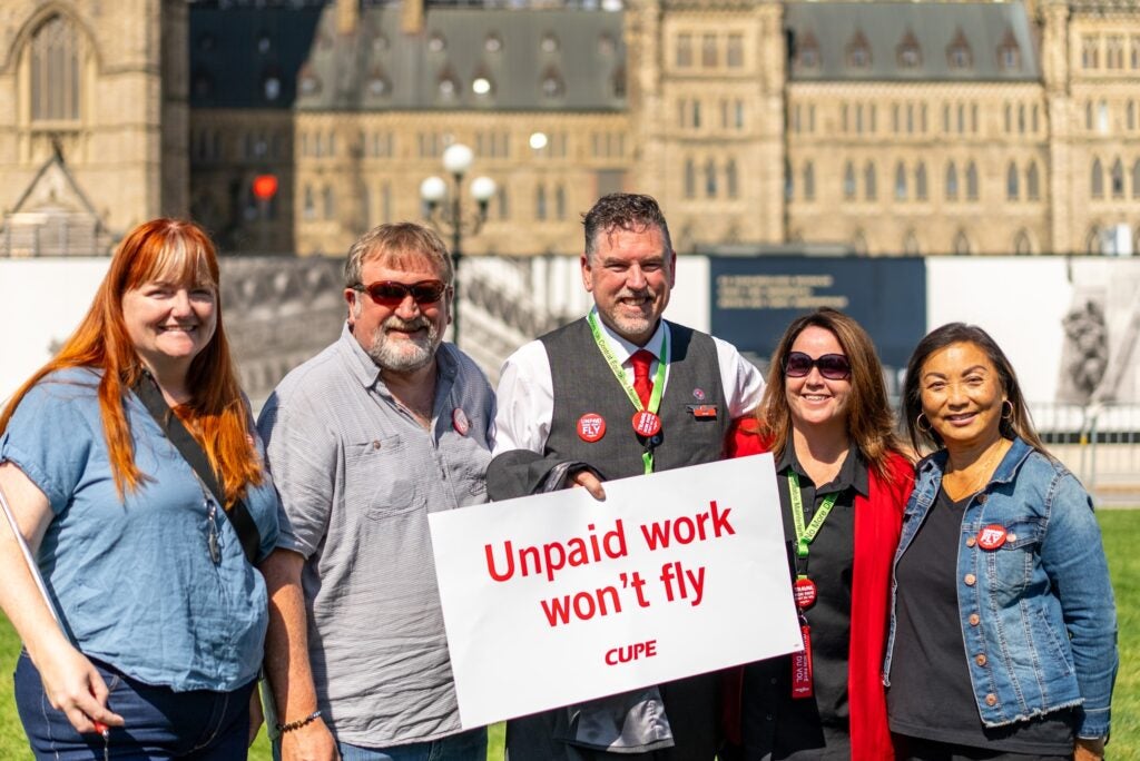 Multiple labour leaders holding an "Unpaid work won't fly" sign in front of Parliament Hill