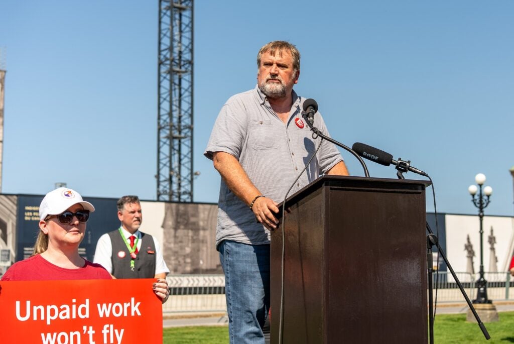 Mark Hancock at a podium in front of Parliament Hill, with a supporter holding an "Unpaid work won't fly" sign